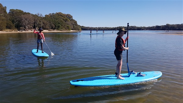 students participating in stand up paddle boarding activity on calm water during outdoor school excursion
